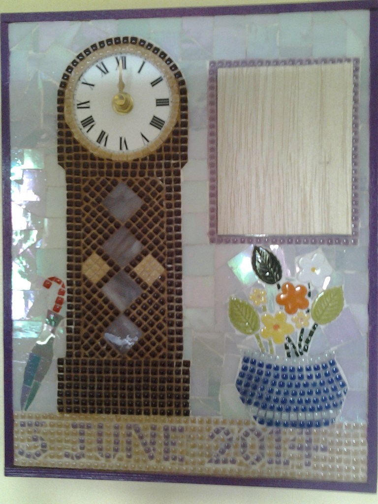 Mosaic depicting a grandfather clock beside which hangs a portrait featuring a photo of the couple on their wedding day. On either side of the clock sits and umbrella for rainy days and a large vase of flowers. These 3 items sit on mosaic lettering showing the date the couple were married.