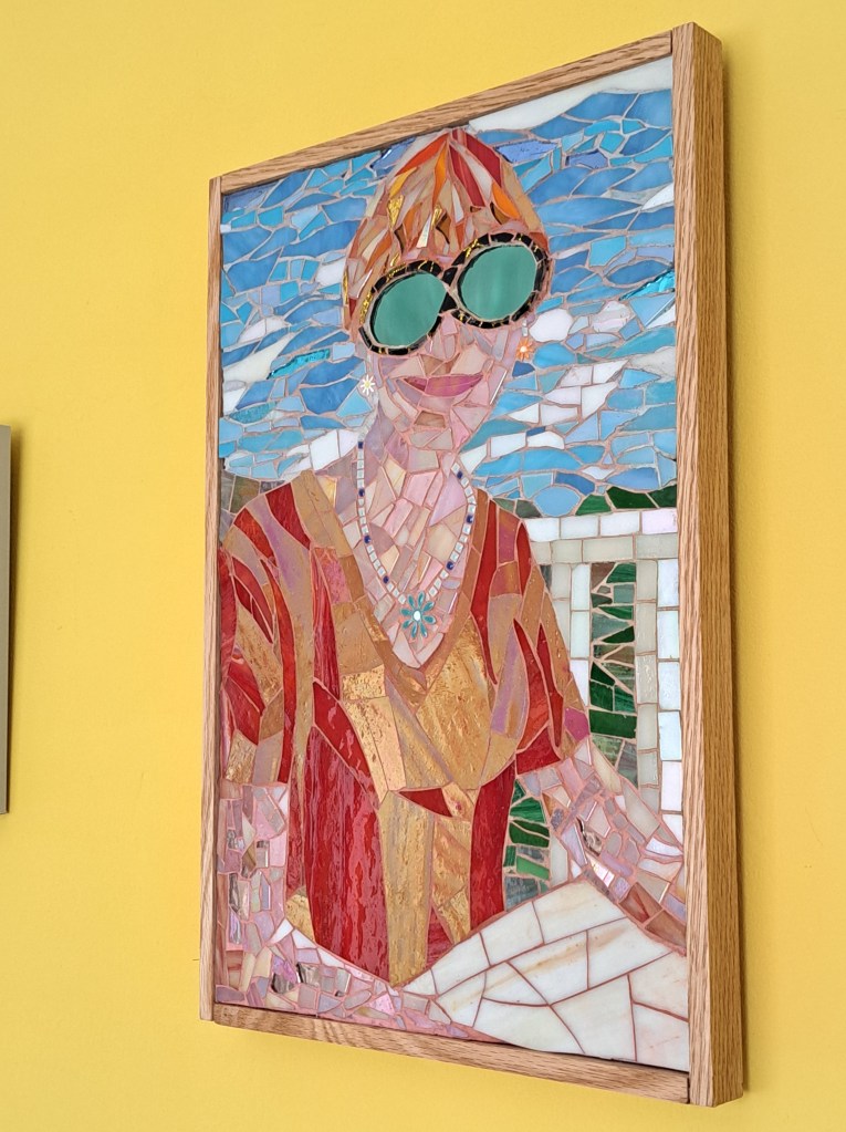 Title: Shades of Green. A mosaic depicting a woman with blonde-red hair sitting at a table and looking at the viewer. She wears a red t-shirt. In the background is a fence and beyond that, a blue sky and green hills.